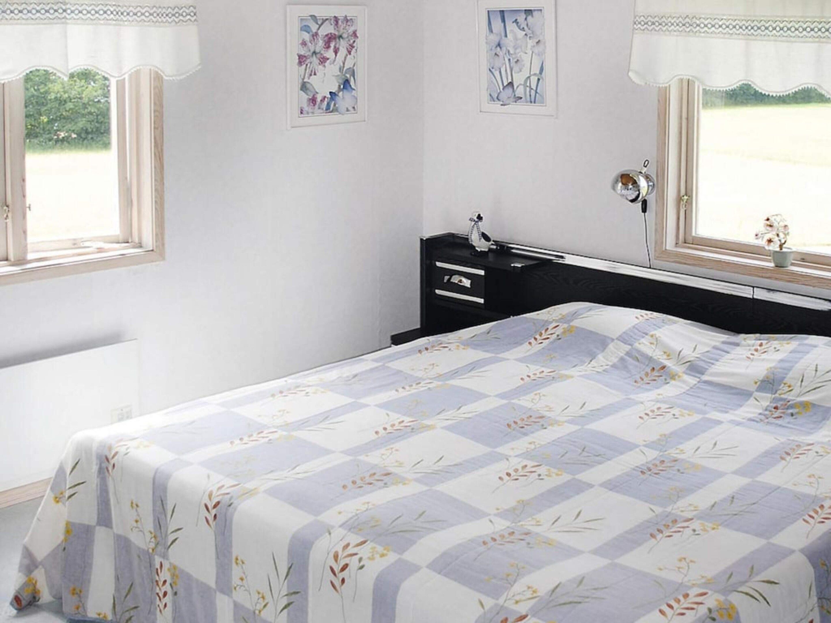 Un dormitorio con cama, mesita de noche, cómoda y dos ventanas con cortinas.