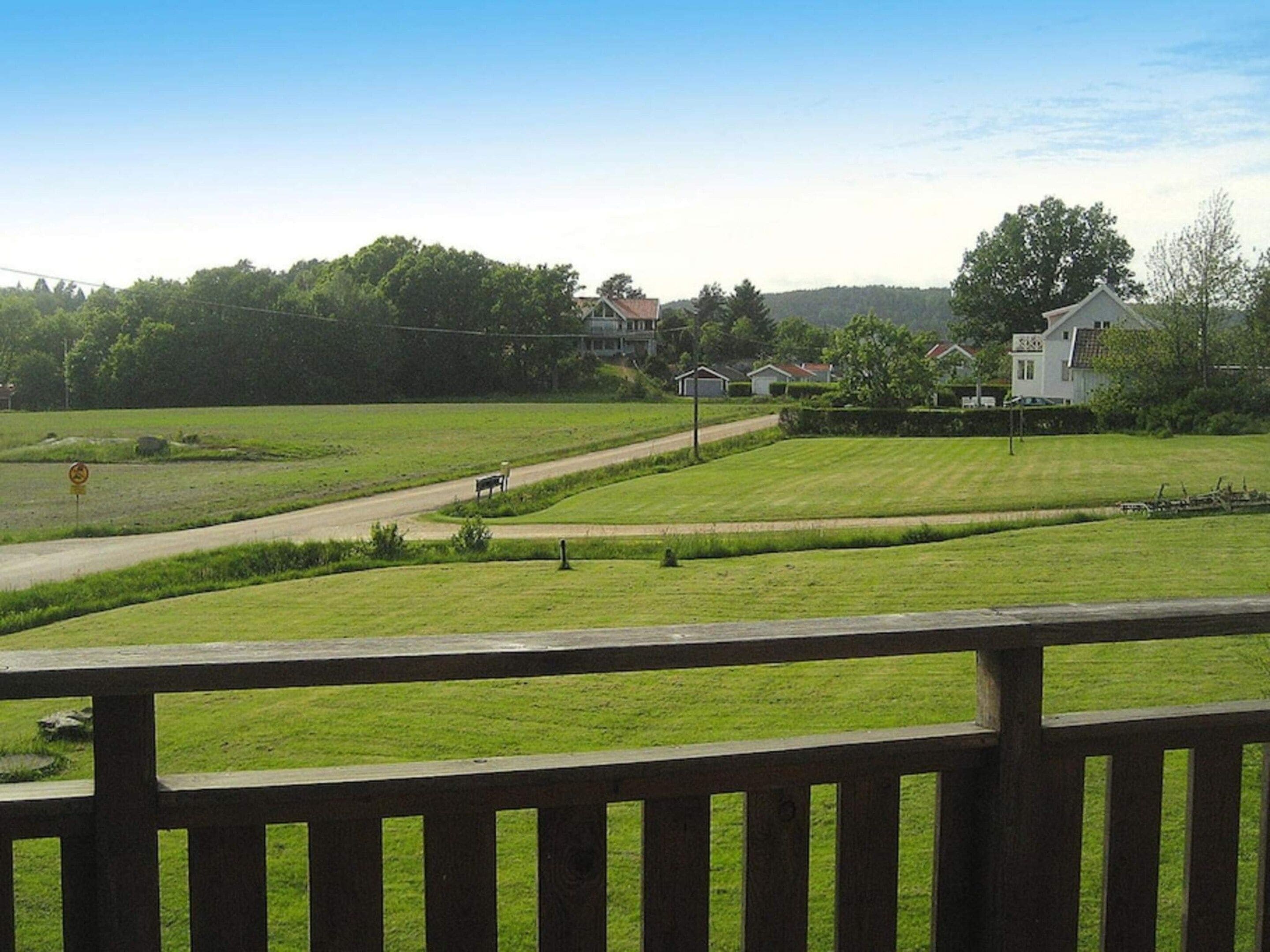 A view from a wooden deck overlooking a grassy area with a road, a bench, and houses in the background.