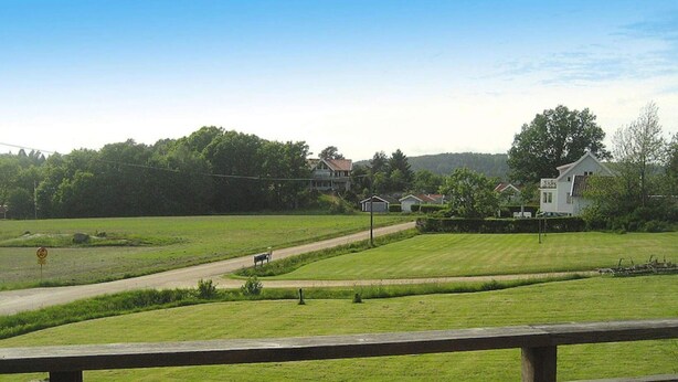 A view from a wooden deck overlooking a grassy area with a road, a bench, and houses in the background. - 4 Person Holiday Home in Henan (Henån)