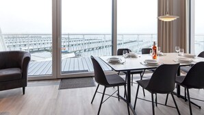 A modern dining area with a large table set for four, large windows with a view of a marina, and a hanging light fixture.