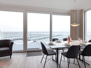 A modern dining area with a large table set for four, large windows with a view of a marina, and a hanging light fixture.