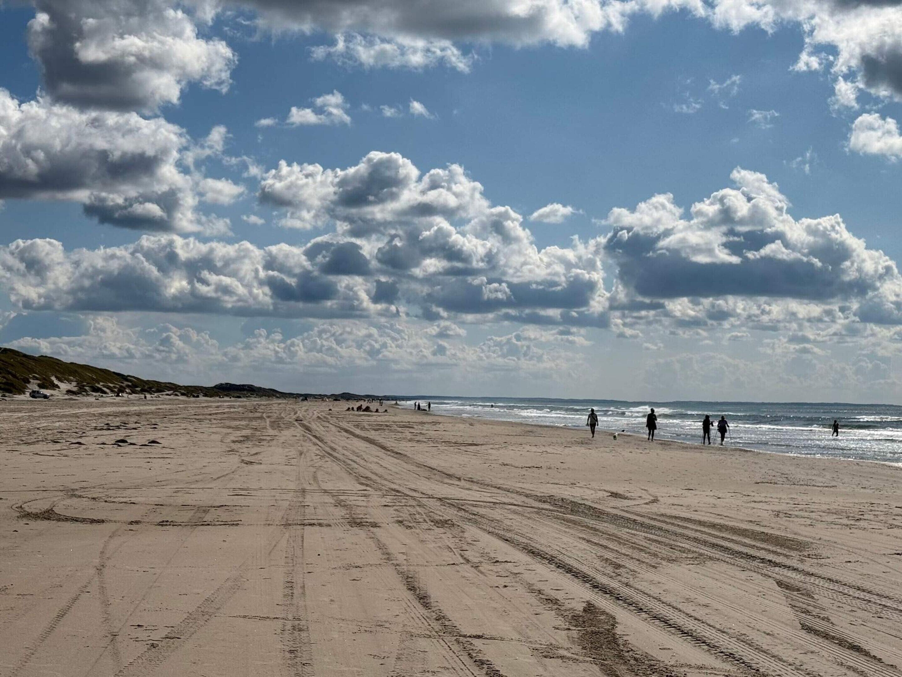 Een breed zandstrand met sporen van autobanden, een heldere lucht met wolken, en mensen die langs de kust wandelen.