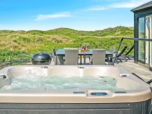 A hot tub with a view of green hills, a table with chairs, and a glass-enclosed area. - 5 Star Holiday Home in Lokken (Lokken)