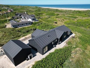 A modern building with a flat roof, surrounded by greenery and a beach in the background.