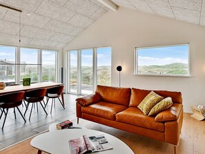 Un salon moderne avec un canapé en cuir, une table à manger, des chaises et de grandes fenêtres donnant sur la campagne.