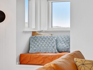 A room with a window, a sofa with patterned cushions, and a brown leather ottoman.