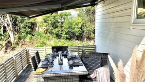 An outdoor dining area with a table set for four, surrounded by wicker furniture, on a wooden deck with a view of greenery and trees. - 4 Star Holiday Home in Halmstad (Halmstad)