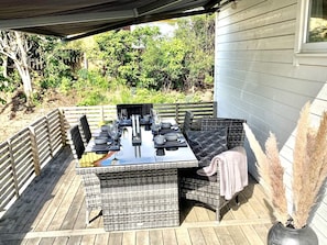 An outdoor dining area with a table set for four, surrounded by wicker furniture, on a wooden deck with a view of greenery and trees. - 4 Star Holiday Home in Halmstad (Halmstad)