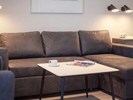 Una sala de estar moderna con un sofá gris, una mesita blanca con una taza y una revista, y dos lámparas de pared.