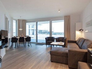 A modern living room with a sofa, a dining area with chairs and a table, and large windows offering a view of the beach. - 5 Star Holiday Home in Wendtorf (Wendtorf)