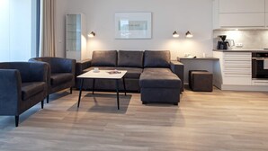 Un salon moderne comprenant un canapé, des fauteuils, une table basse et un coin cuisine avec des placards et des appareils électroménagers.