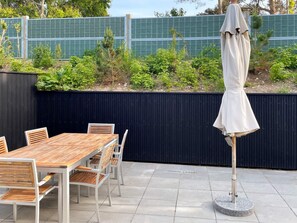 An outdoor patio with a wooden table and chairs, a metal umbrella, and a black fence with greenery behind it.