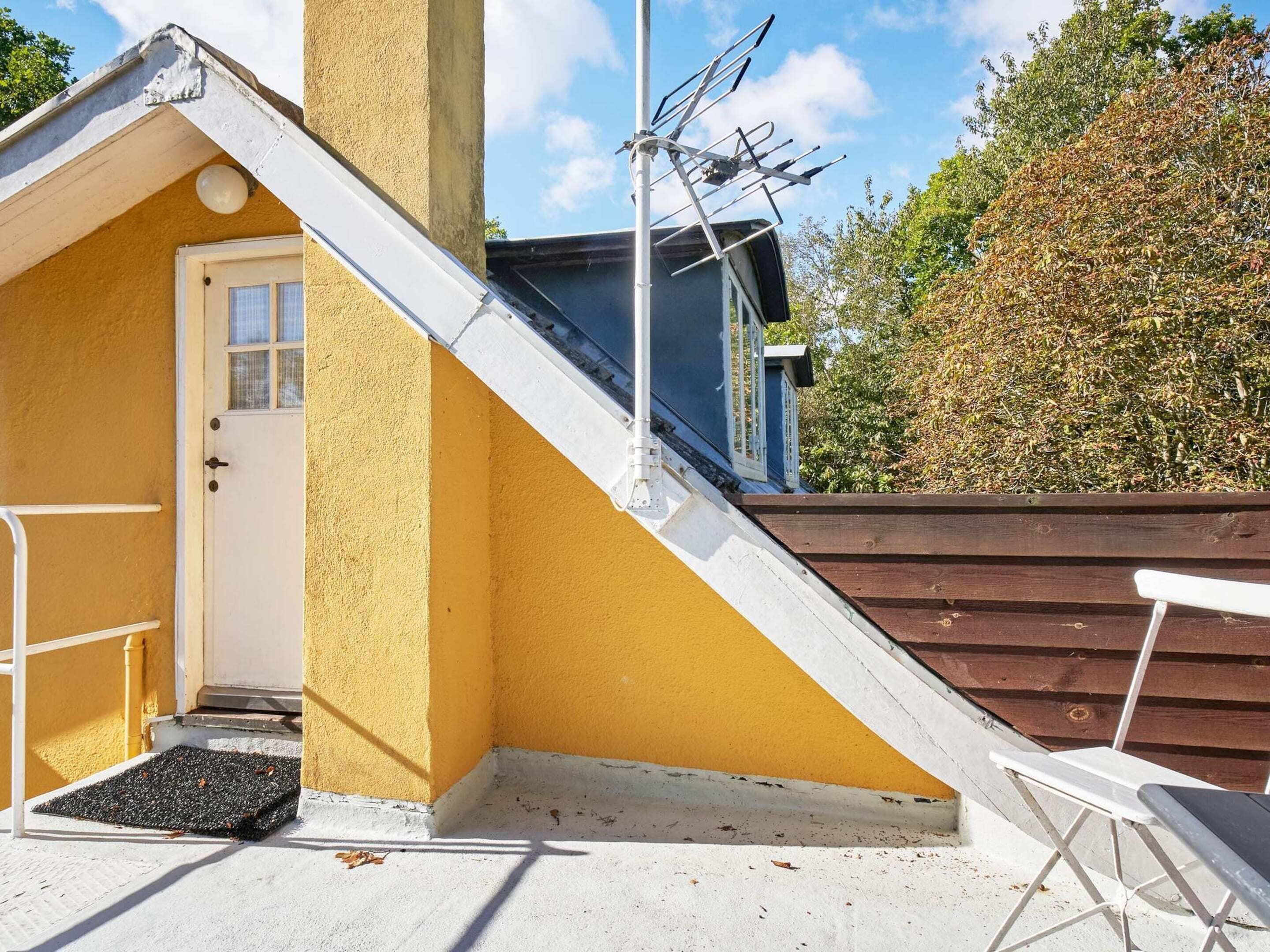 Un pequeño edificio amarillo con una puerta blanca, un banco de madera y una silla de metal en el exterior.