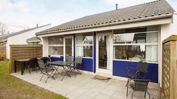 A single-story house with a patio area featuring a table and chairs, large windows, and a tiled roof.