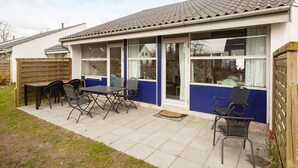 A single-story house with a patio area, a dining table with chairs, and a window with curtains. - 4 Star Holiday Home in Vaeggerlose (Vaeggerlose)