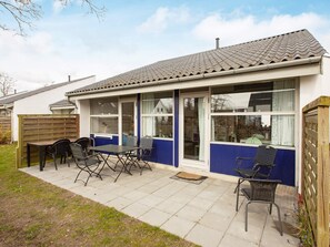 A single-story house with a patio area, a dining table with chairs, and a window with curtains. - 4 Star Holiday Home in Vaeggerlose (Vaeggerlose)