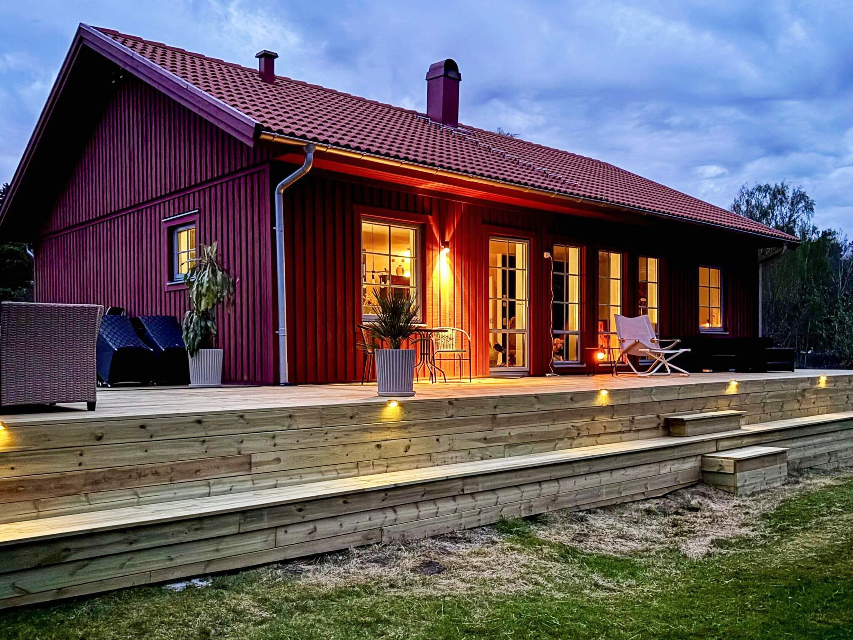 Een rode houten woning met schuin dak, grote ramen en een houten terras met buitenmeubels.