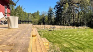 A wooden deck with a red building in the background, a grassy lawn, and a tennis court. - 5 Star Holiday Home in Klovedal (Klovedal)