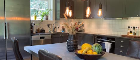 A modern kitchen with a stainless steel refrigerator, a dining table with chairs, a fruit bowl, and a vase with dried plants.