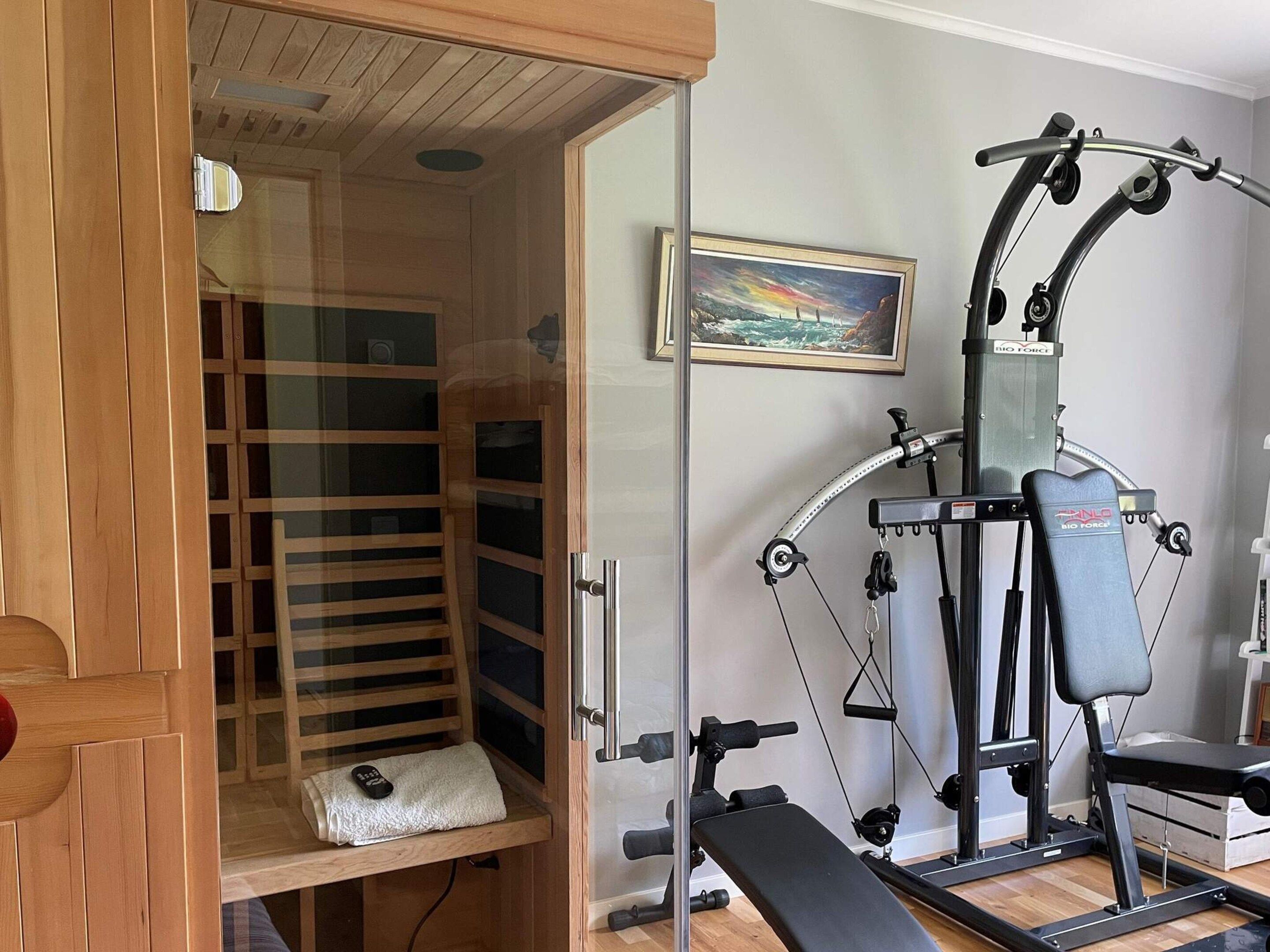 Eine Sauna mit einer Bank und einer Glastür, einem an der Wand befestigten Trainingsgerät und einem gerahmten Gemälde.