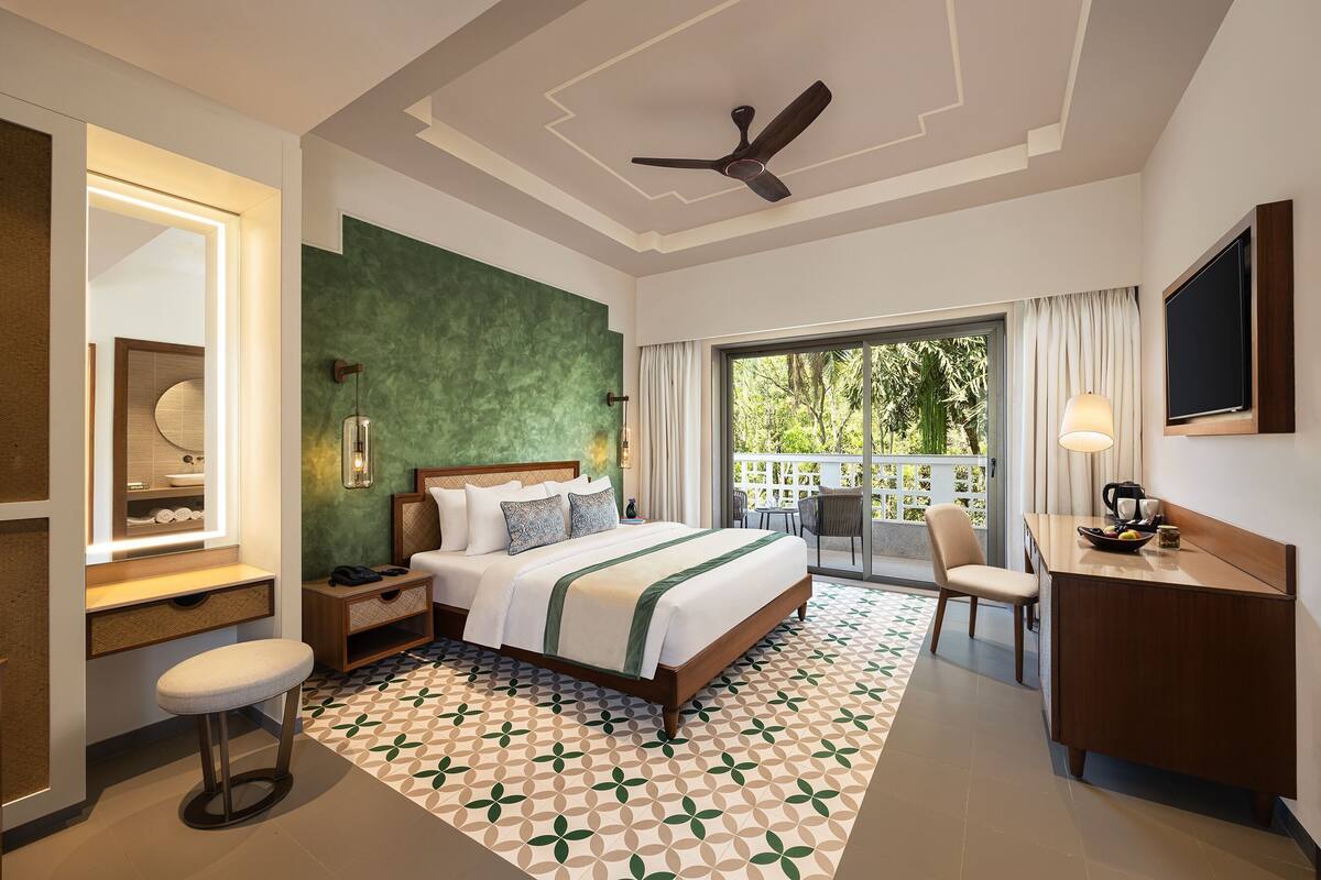 A hotel room with a bed, a desk, a chair, a TV, a ceiling fan, and a balcony with a view.