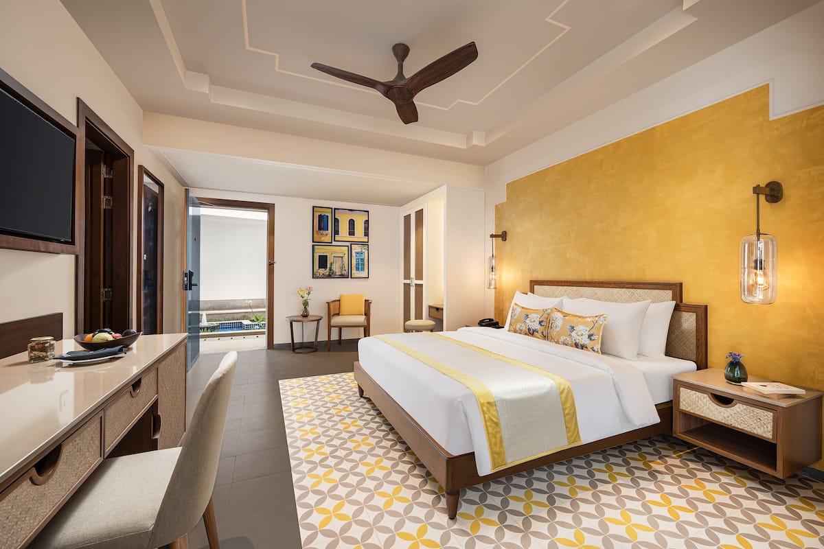 A hotel room with a bed, bedside table, and a desk with a chair. There is a ceiling fan, a window, and a door leading to another room.