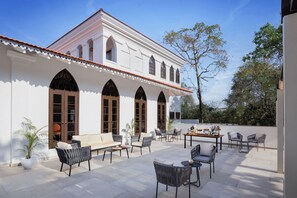 A courtyard with outdoor seating, a white building with arched windows, and a tree. - The Yellow House, Goa – IHCL SeleQtions (Anjuna)