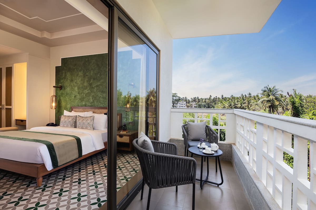 A hotel room with a bed, a balcony with a view of palm trees, and a dining area with chairs and a table.