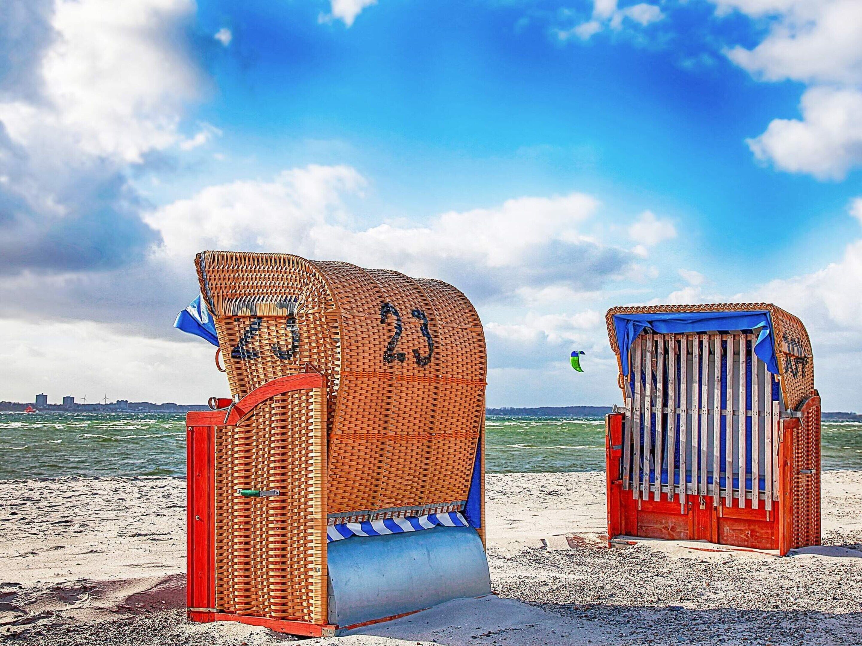 Zwei Strandkörbe mit den Nummern 23 und 24, ein Sandstrand und im Hintergrund ein Gewässer.