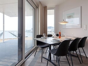 Espaço de refeições com vista para o mar, mesa posta com pratos e copos, e uma garrafa de vinho.