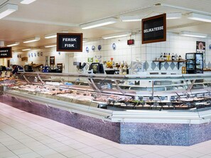 A deli counter with various meats and seafood, overhead signs indicating 'FERSK FISK' and 'DELIKATESSE', and a tiled wall with a blue and white pattern.