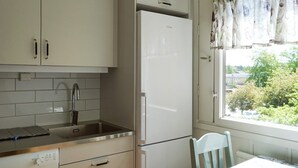 Cozinha com armários brancos, pia de aço inoxidável e geladeira. Há uma janela com vista para o verde lá fora.