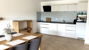 A modern kitchen with white cabinets, a wooden dining table with chairs, and a large window.