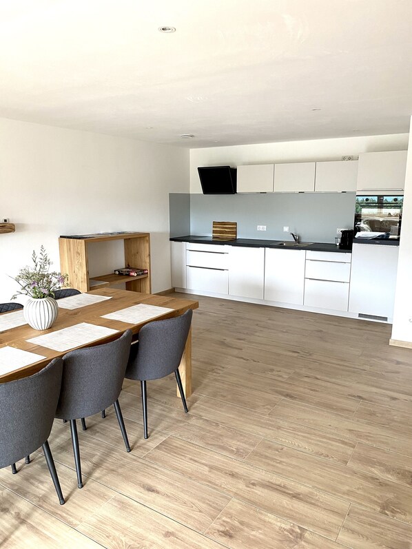 A modern kitchen with white cabinets, a wooden dining table with chairs, and a large window. - DasFranz 2 (Weitnau)