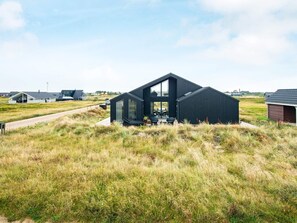 A modern black building with a large glass window and a flat roof, surrounded by grassy fields and other buildings in the background.