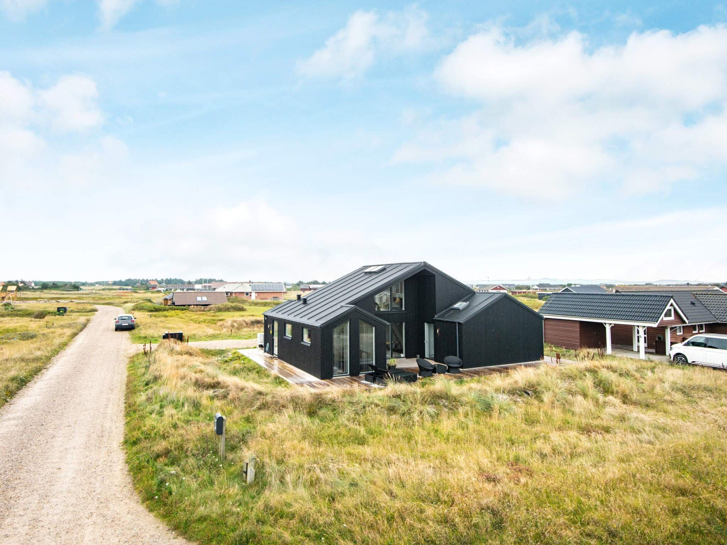 Et moderne, sort hus med skråt tag, omgivet af et landligt landskab med en grusvej, der fører hen til det.