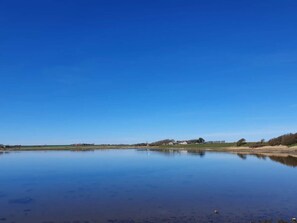 A calm lake with a clear sky, surrounded by open fields and a few buildings in the distance. - 10 Person Holiday Home in Karby (Øster Assels)