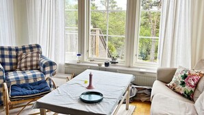 Una sala de estar acogedora con un sofá escocés, un sillón estampado con flores, una mesita con un plato y una vela, y ventanas amplias con vistas a árboles.