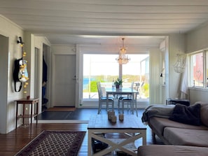 A room with a wooden floor, a sofa, a coffee table, a dining table with chairs, a chandelier, and a view through a glass door.