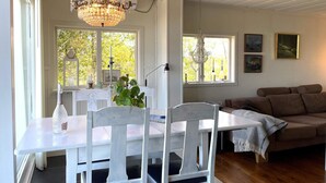 A dining area with a white table, chairs, a chandelier, and a view of trees through large windows.