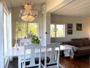 A dining area with a white table, chairs, a chandelier, and a view of trees through large windows.