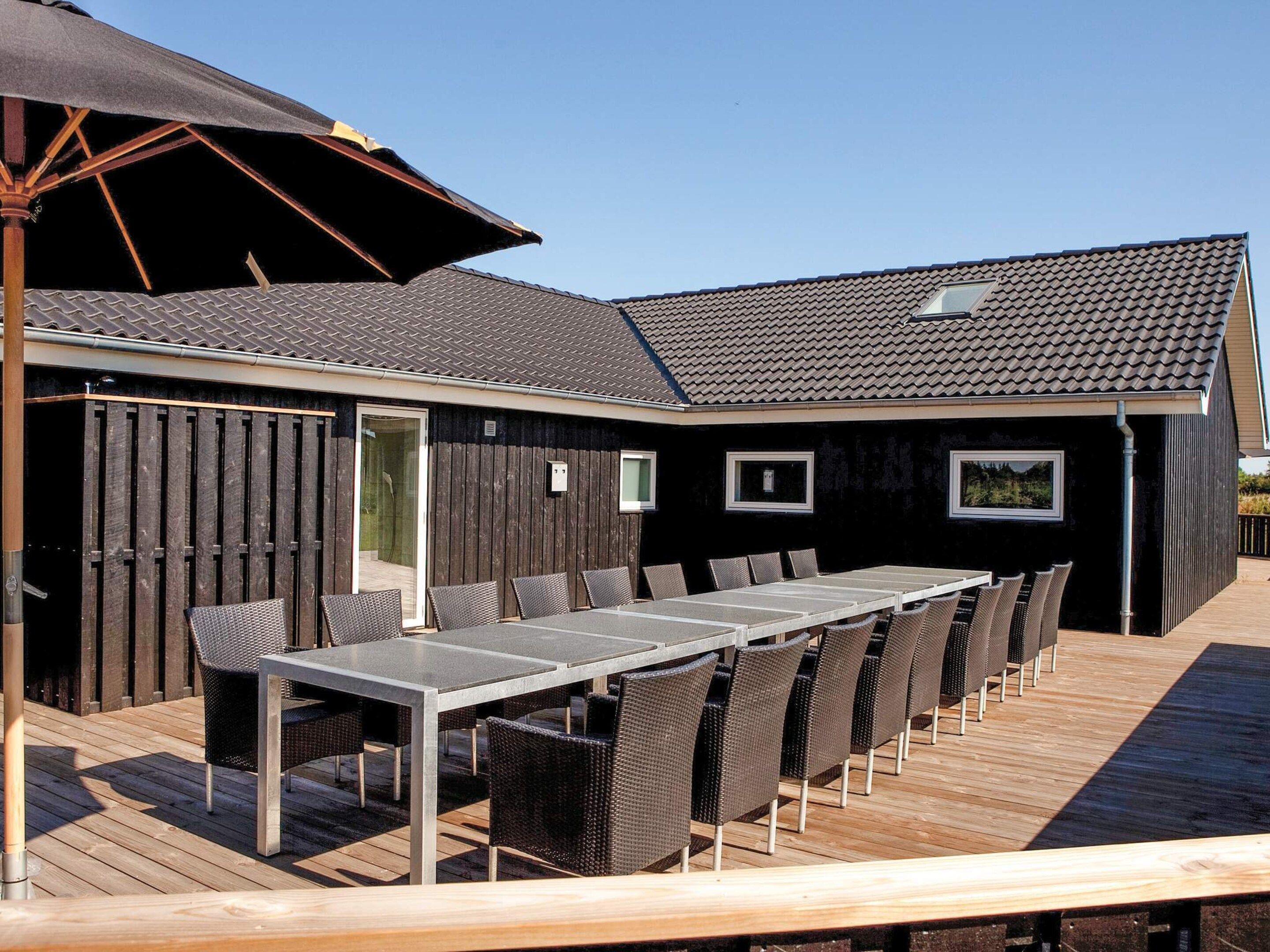 An outdoor deck with a long table and chairs, a wooden fence, and a building with a tiled roof.