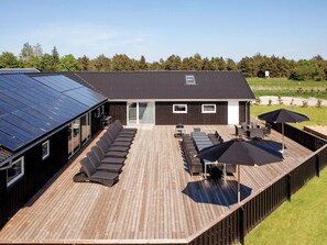 A deck with sun loungers and umbrellas, a barbecue area, and a building with a solar panel roof.