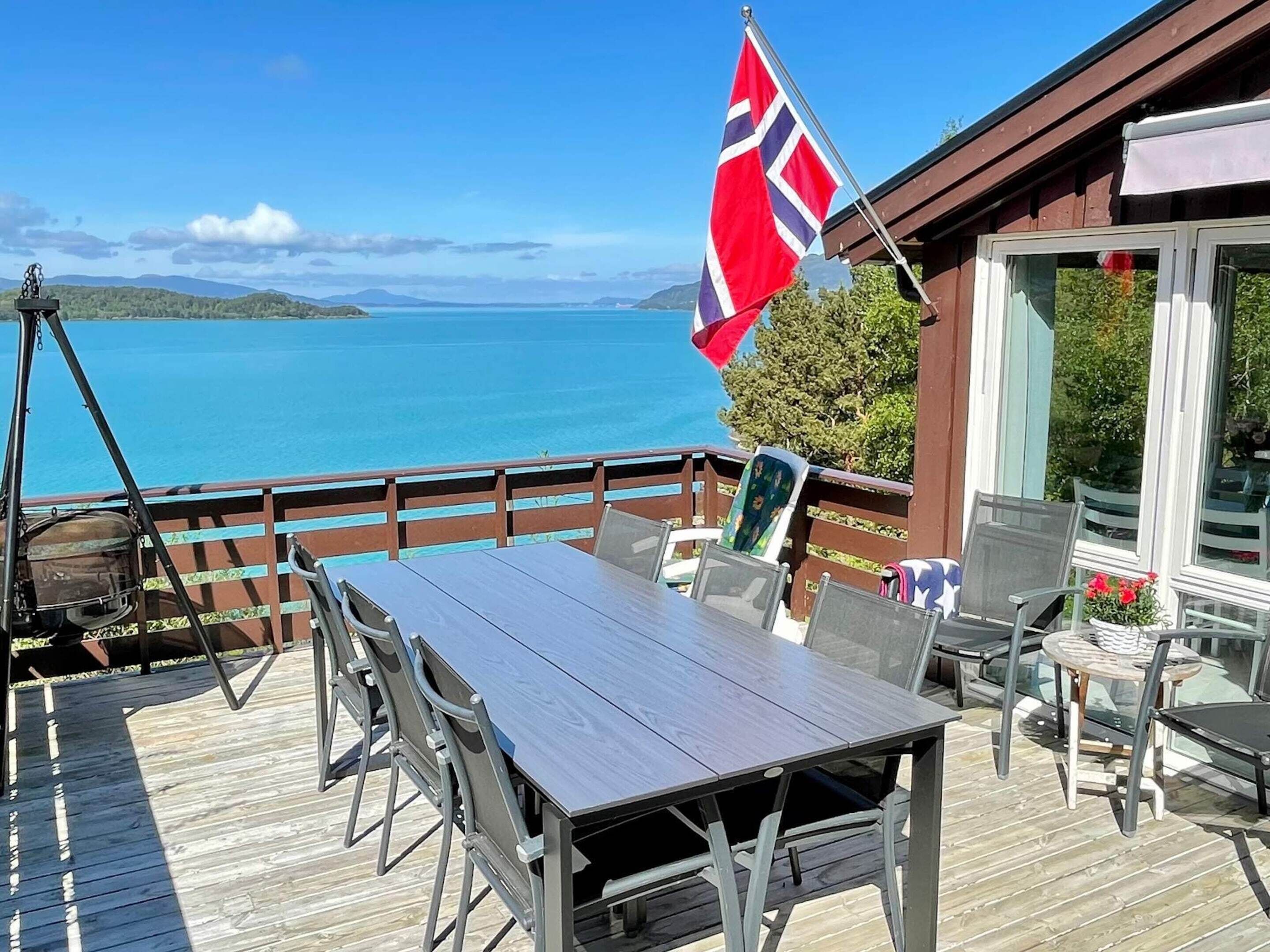 A wooden deck with a table and chairs, overlooking a body of water and hills.