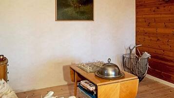 A room with a wooden floor, a small wooden table with a metal container, a framed picture on the wall, and a metal basket with items.