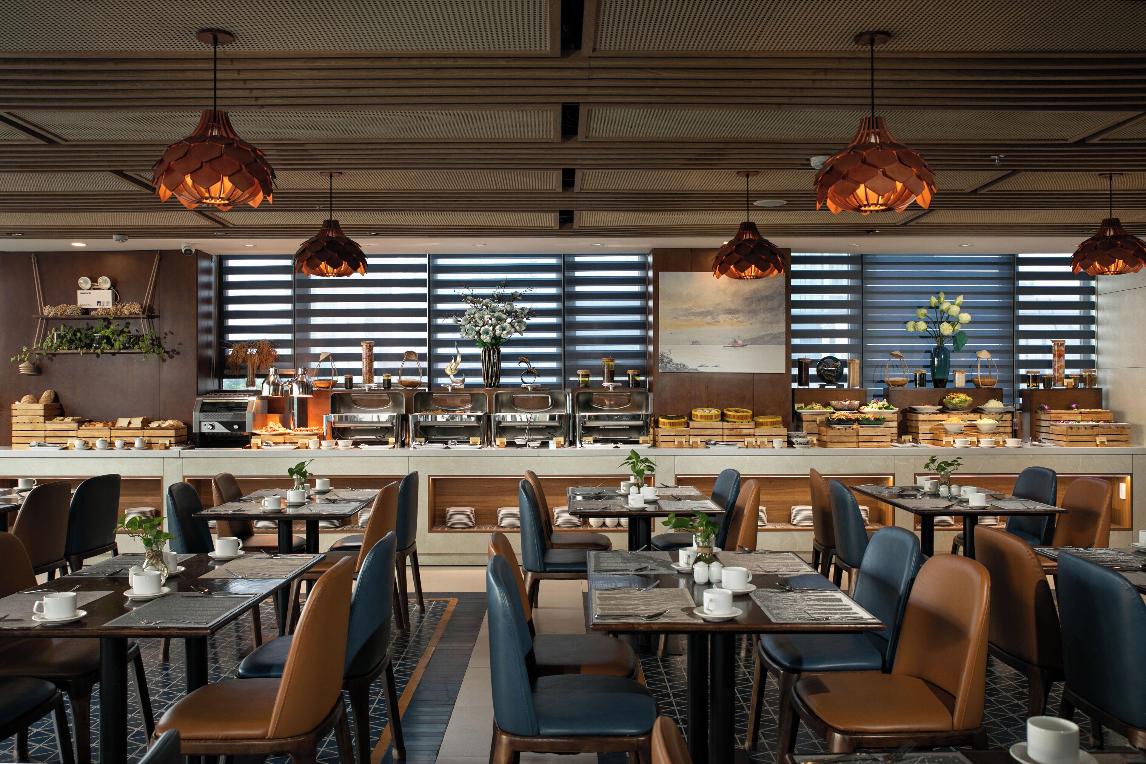 A modern restaurant with multiple dining tables and chairs, large windows with blinds, and hanging pendant lights.