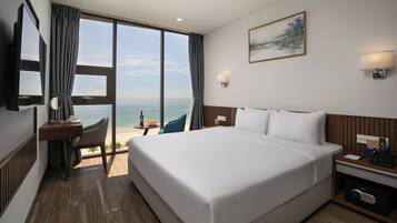 A hotel room with a large bed, a desk with a chair, a TV, and a view of the ocean.