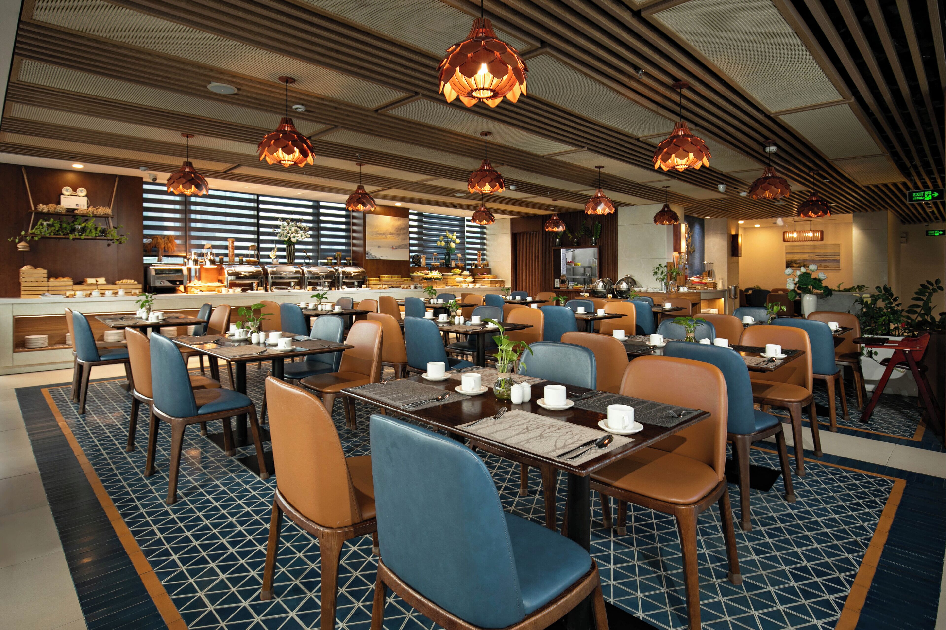 A hotel dining area with multiple tables and chairs, overhead lighting, and a kitchen in the background.