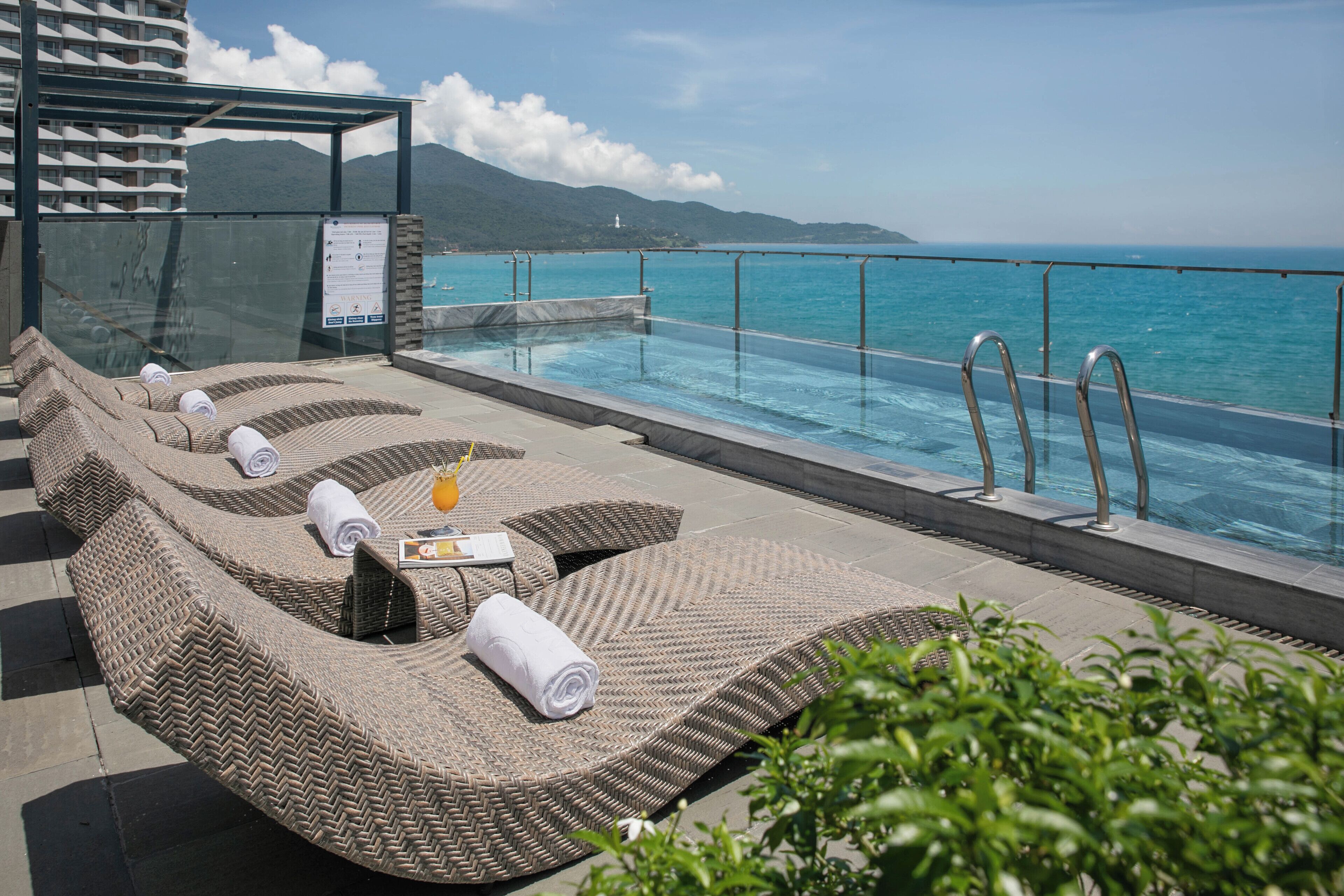 A rooftop infinity pool with wicker loungers, a glass railing, and a view of the sea and mountains.
