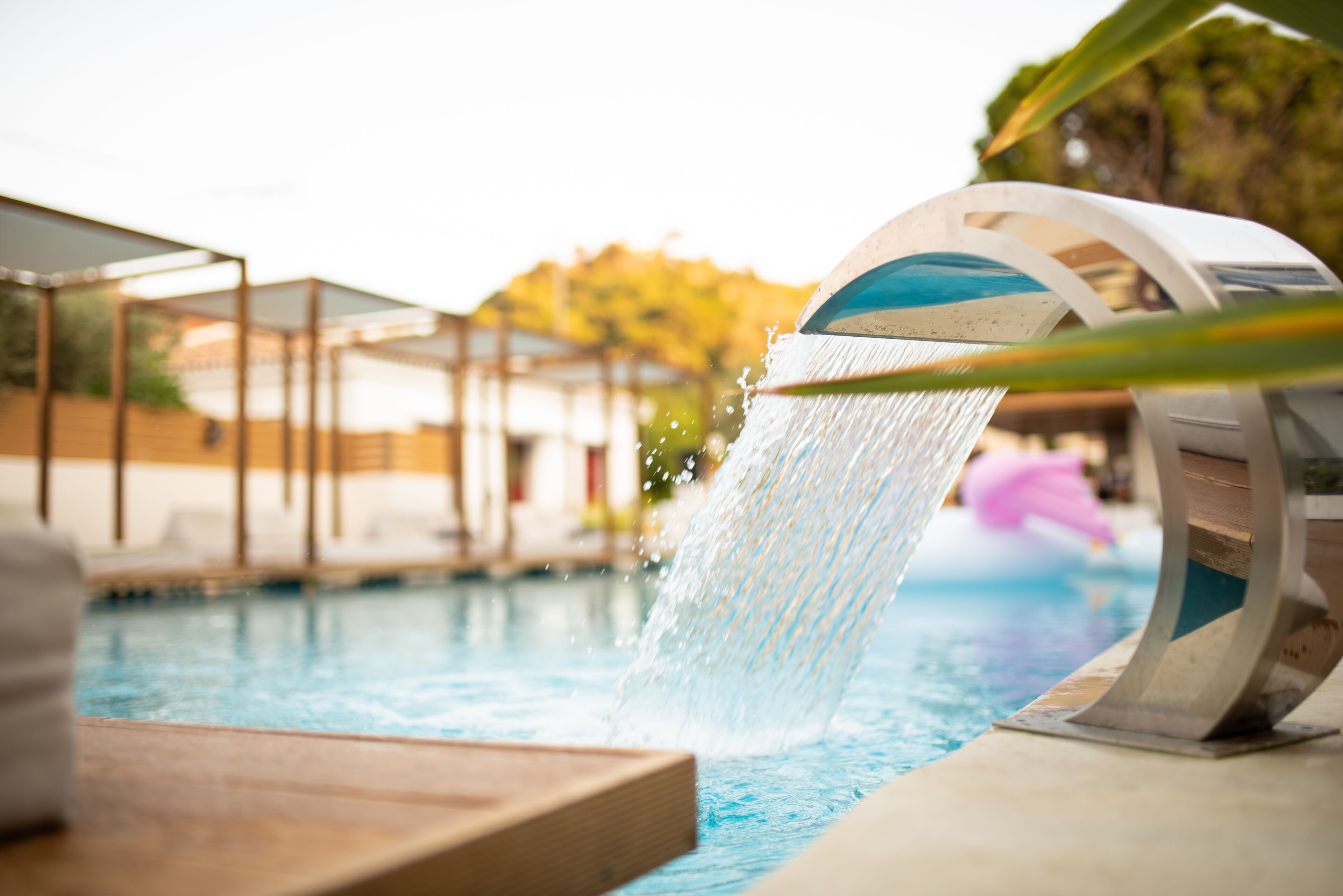 Ein Pool mit Wasserspiel, einer Holzterrasse und einem überdachten Sitzbereich.